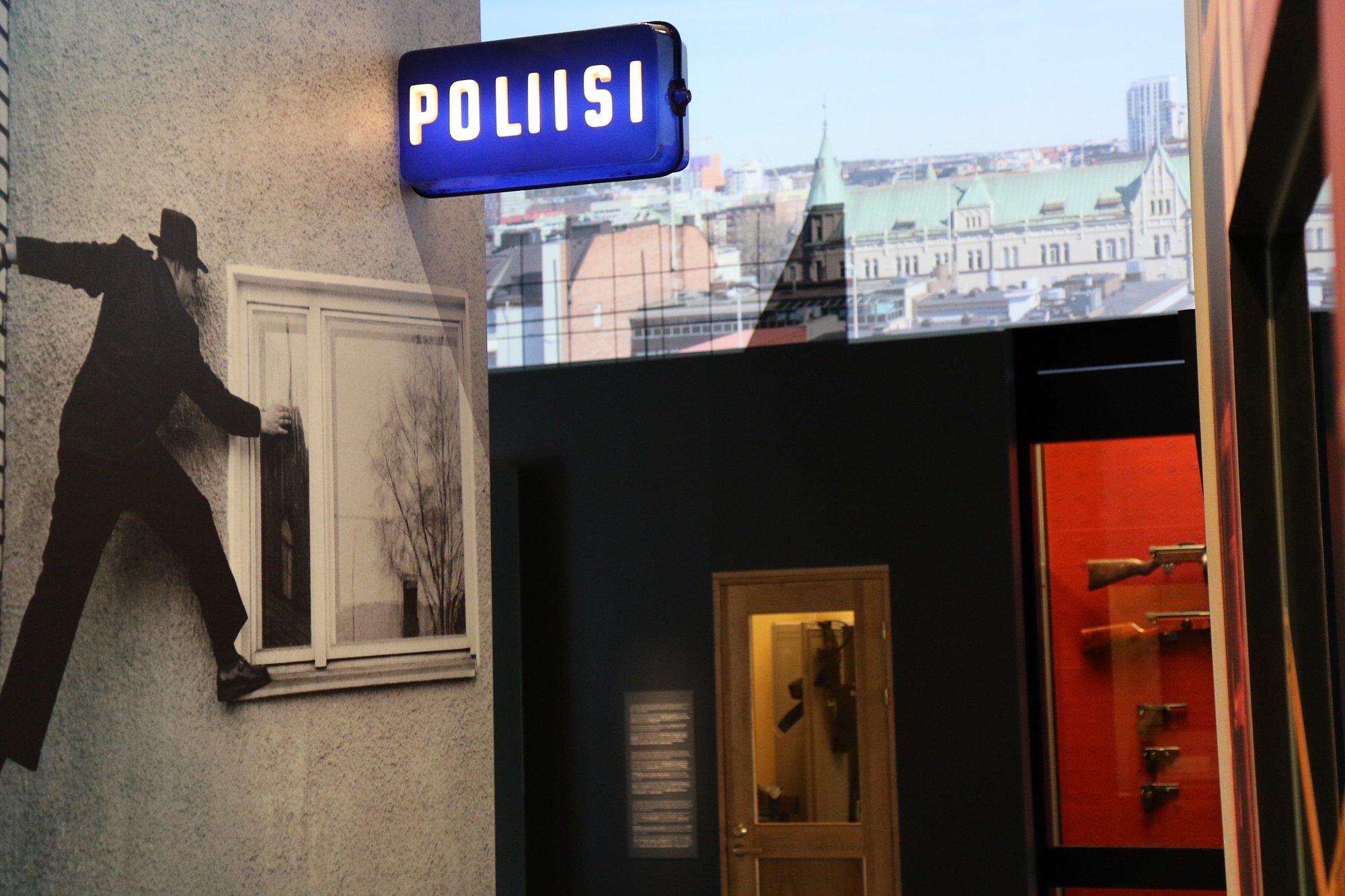 Bild av museets utställning. På bilden finns bland annat en polisskylt, vapen och på väggen ett stort svartvitt fotografi på vilket en man klättrar längs ytterväggen på ett hus med ett ben på fönsterbrädet. Bild Polismuseet, Jarkko Järvinen.